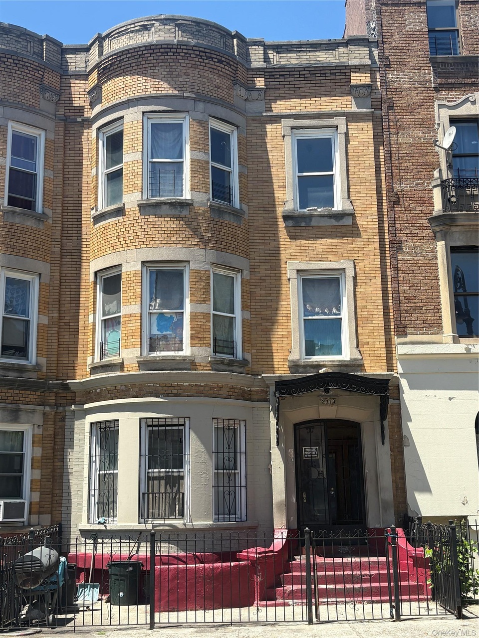 Street view of 2313 Newkirk Avenue, a three-story tan and brown brick row house in Ditmas Park, Brooklyn, with a rounded bay window, security bars on the lower windows, and red plywood covering the front stoop ahead of renovation